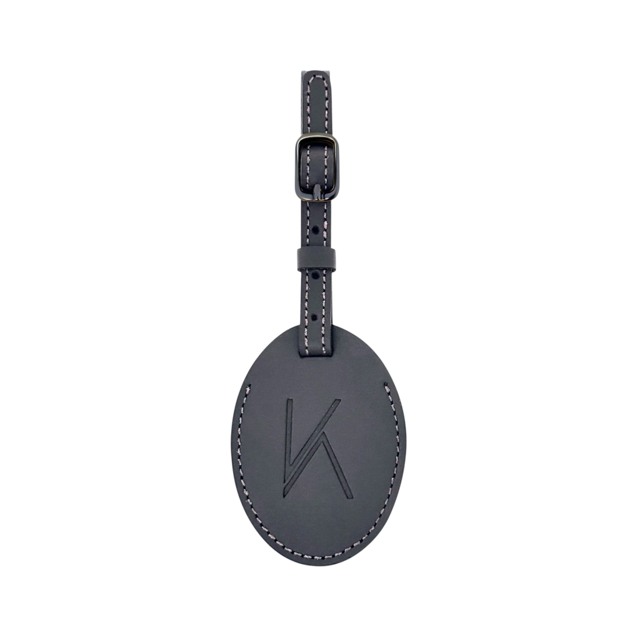 A black oval leather luggage tag with a matching strap and buckle, featuring an embossed stylized 'K' logo on the front, shown against a plain white background.